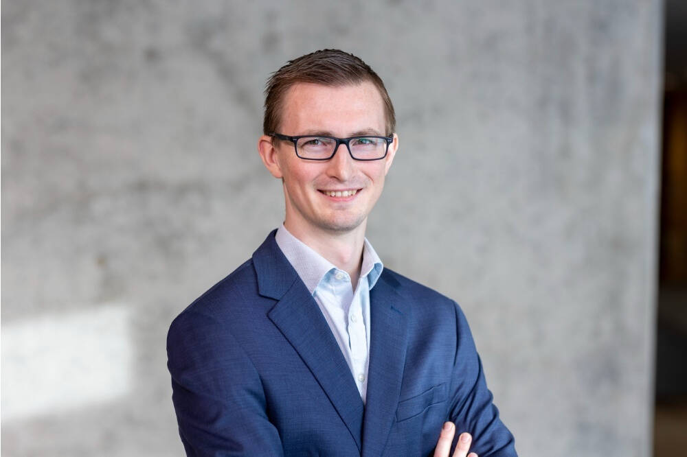A man wearing a blue suit smiles at a camera. He is wearing glasses and is standing in front of  a gray background